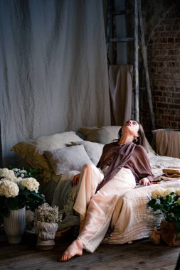 Yatakta pozlar veren güzel, genç bir kadın. İç döşemesi kaliteli. Etrafta bir sürü çiçek var. Arka planda tavan arası tipi tuğla duvar.