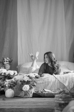 Yatakta pozlar veren güzel, genç bir kadın. İç döşemesi kaliteli. Etrafta bir sürü çiçek var. Arka planda tavan arası tipi tuğla duvar.