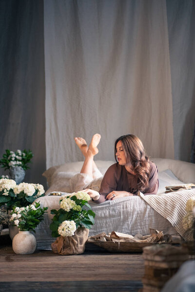 Beautiful young woman posing on bed in room with vintage interior, a lot of flowers around. Loft style brick wall in background.