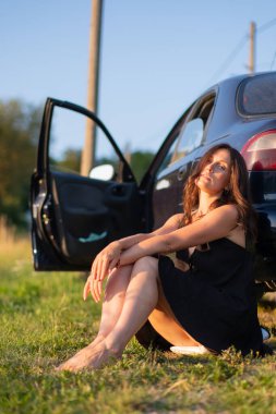 Siyah elbiseli bir kız eski bir arabanın yanında güzel poz verir. Güneş, eski bir arabanın yanında poz veren güzel kızın yüzünde parlıyor..