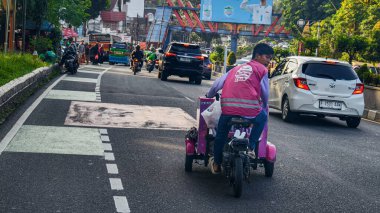Bogor, Endonezya - 3 Eylül 2025: Meşgul bir Asya caddesinde şehir trafiğinde mal teslimatı yapan pembe yelekli motorlu trishaw sürücüsü.