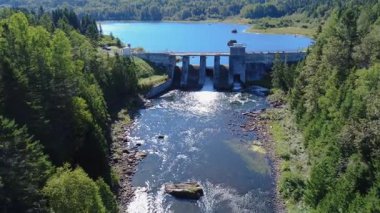 Panoramik hava manzarası bir göle ve beton yapısının içinden geçen eski bir hidroelektrik barajına bakıyor. Mitis Barajı, Göl & Mitis Nehri, Quebec, Kanada, 2025.