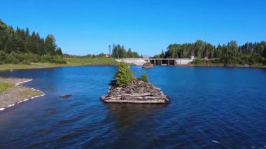 Eski kaynakların kalıntılarıyla dolu bir orman gölü ve açık mavi gökyüzünün altında bir baraj içeren huzurlu doğal bir manzara. Mitis Barajı, Lac-des-Eaux-Mortes, Quebec, Kanada, 2025.