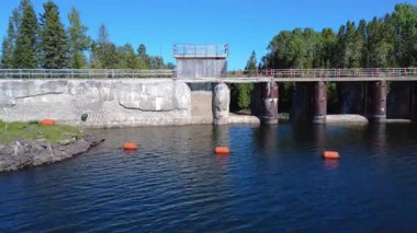 Eski bir barajın çelik ve beton yapısının içindeki insansız hava aracı uçuşu, berrak mavi gökyüzünün altında canlı yeşil ağaçlarla çevrili sakin bir nehri ortaya çıkarıyor. Mitis Barajı, Quebec, Kanada, 2025.