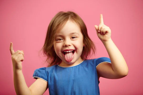 Niña alegre bailando con las manos levantadas y la lengua sobresaliendo, retrato de niña alegre sobre fondo rosa . — Foto de Stock