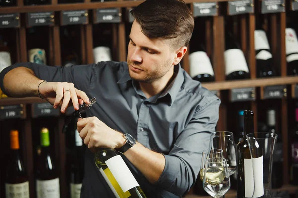 Sommelier opens with corkscrew bottle of white wine, next to the decanter and glasses, on wine shelf background in cellar.