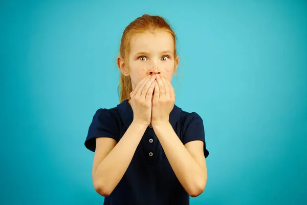 Rothaarige Mädchen mit großen Augen der Angst bedeckt Mund mit Händen vor blauem Hintergrund. — Stockfoto