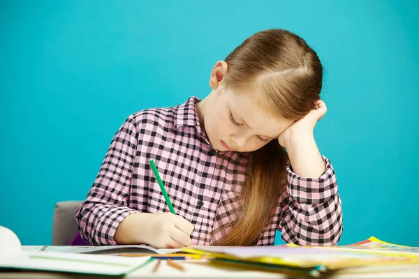 El plano horizontal de la muchacha en el escritorio escribe cuidadosamente en la tarea del cuaderno, sentado sobre fondo azul aislado. Niño realiza la tarea . — Foto de Stock