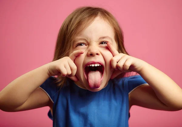 Retrato de menina grimace e mostra a língua no rosa isolado — Fotografia de Stock