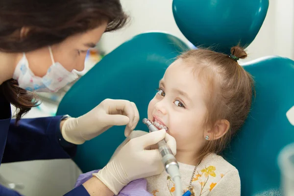 Professional dentist in facial mask polishes childs teeth with modern electric tool. Kid has no fear during medical procedure of teeth grinding in modern office.