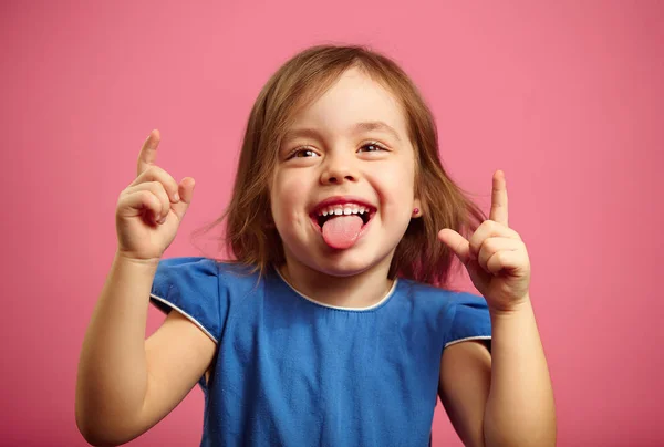 Веселая маленькая девочка танцует с поднятыми руками и торчащим языком, портрет веселой девочки на розовом фоне . — стоковое фото