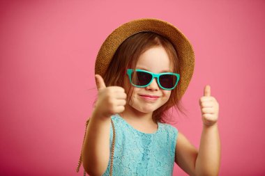 Neşeli kız plaj şapka ve güneş gözlüğü, başparmak geliyor, iyi bir ruh hali var, izole pembe standları.