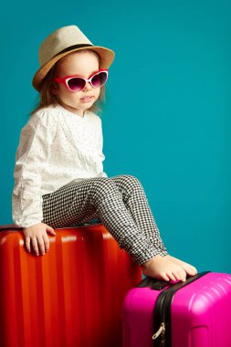 Üzücü bir kötü ruh hali, portre izole mavi valiz, hasır şapka ve güneş gözlüğü, hayal kırıklığına uğramış küçük kız oturan çocuktur.
