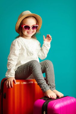 Hasır şapka ve güneş gözlüğü bavul üzerinde oturan bir yolculuk, izole izole mavi üzerinde atış studio oluyor güzel çocuk portresi, neşeli küçük kız.