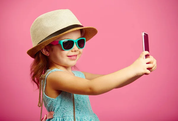Beautiful little girl dressed in summer dress, wearing a straw hat and sunglasses, makes a selfie portrait on the phone, stands over isolated pink. — Stock Photo, Image