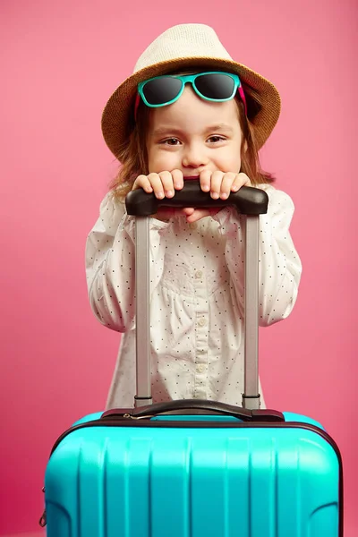 Caucasian little girl in sunglasses and straw hat, holding a suitcase, standing on isolated pink. — Stock Photo, Image