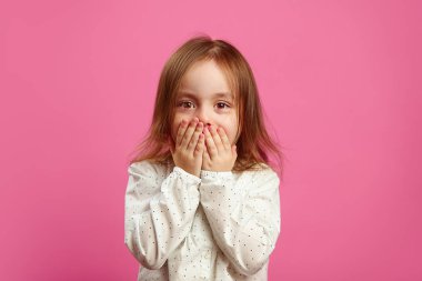 Sürpriz bir görünüm ile küçük kız elleri ile ağzını kaplı ve pembe izole arka planda size bakıyor.