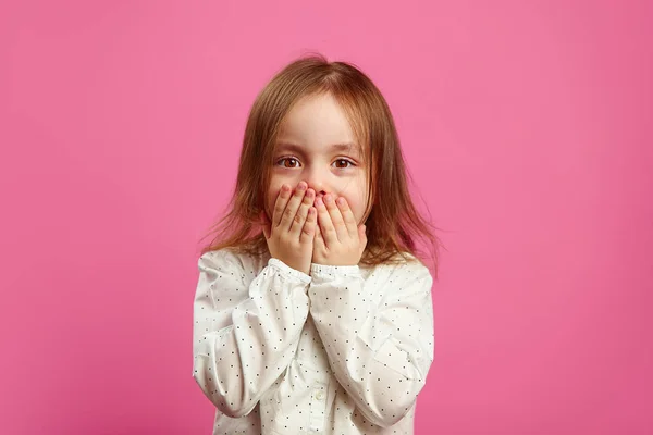 Klein meisje met een verbaasde blik bedekt haar mond met handen en kijkt naar je op roze geïsoleerde achtergrond. — Stockfoto