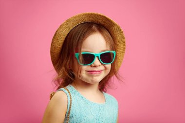 Hasır şapka ve güneş gözlüğü ile güzel küçük kız Portresi, mavi elbise giyer, pembe izole duruyor.