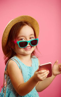 Güzel küçük bir kız, çekici bir gülümseme ile giyinmiş hasır şapka ve güneş gözlüğü, kameraya bakarak, bir telefon tutarak, izole pink ayakta.