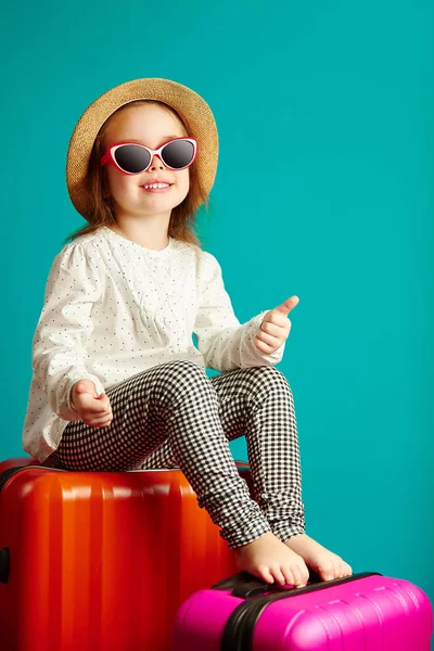 Radosna dziewczynka to będzie podróż, siedzi na walizkach, noszenie słomkowy kapelusz i okulary przeciwsłoneczne, pokazuje kciuk w górę, portret dziecko na na białym tle niebieski. — Zdjęcie stockowe
