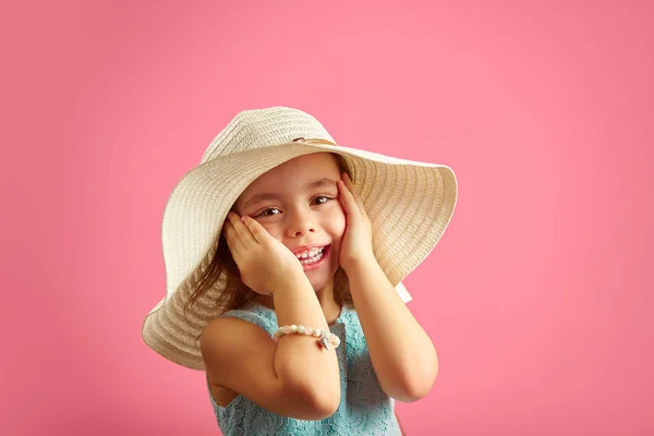 Surprised little girl in straw hat and blue dress, puts her hands to face, expresses surprise, shows positive emotion. Portrait of joyful child on pink isolated background.