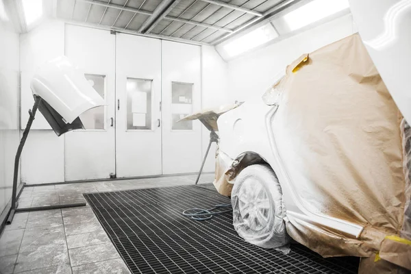Painting the white car body in the spray booth in the body repair workshop, elements are covered with protective paper.