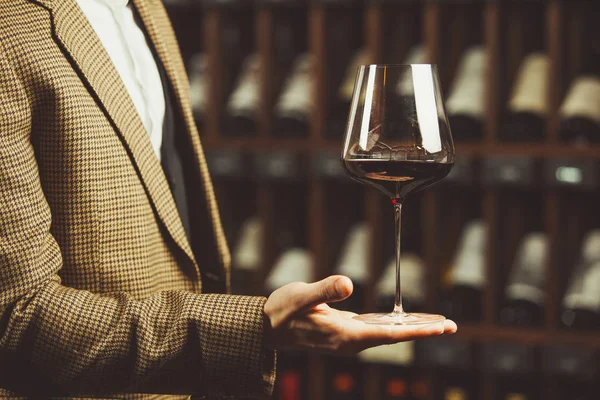 Ποτήρι κόκκινο κρασί στο χέρι sommelier στο κελάρι με φόντο μπουκάλια. — Φωτογραφία Αρχείου