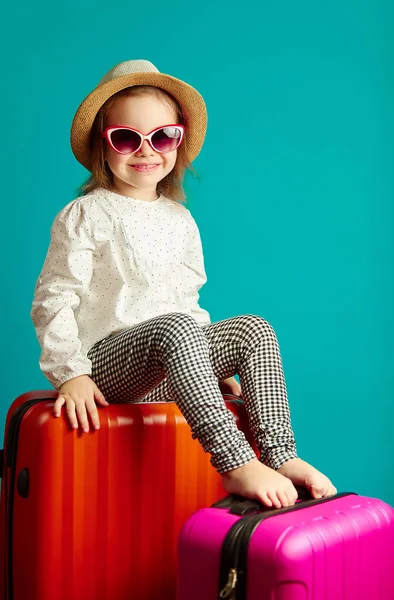 Dziewczynka uśmiechający się w słomkowy kapelusz i okulary przeciwsłoneczne, siedzi na walizkach, portret piękne dziecko wybiera się w podróż, studio strzał na na białym tle niebieski na białym tle. — Zdjęcie stockowe