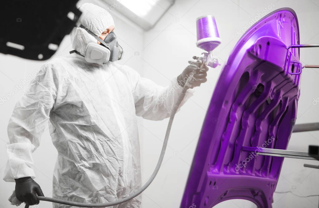Worker paints hood of auto in violet color with spray gun. White car in a paint chamber during repair work.