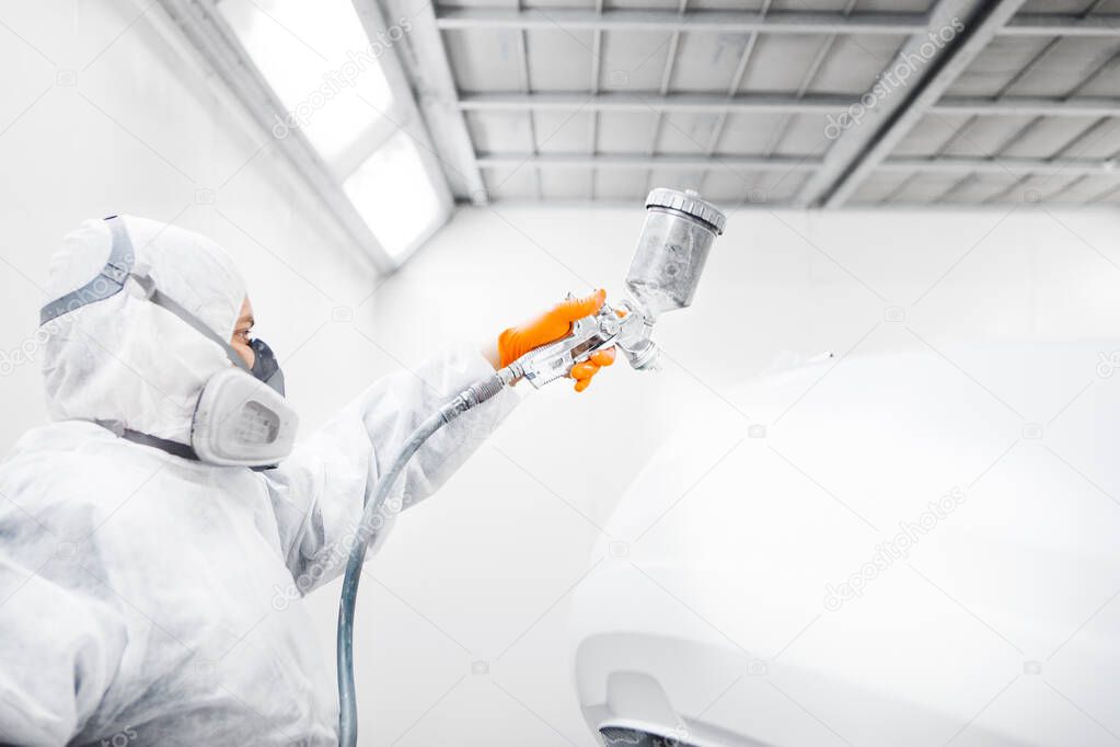 Auto mechanic worker painting a white car with spray gun in a paint chamber during repair work.