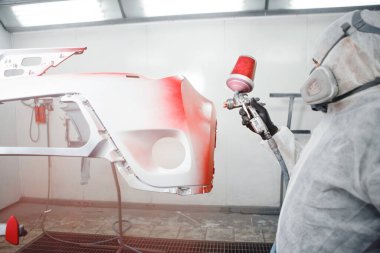 Odamdaki erkek tamirci araba elementlerini kırmızıya boyuyor. İşçi aracı sprey silahla beyaza boyuyor..