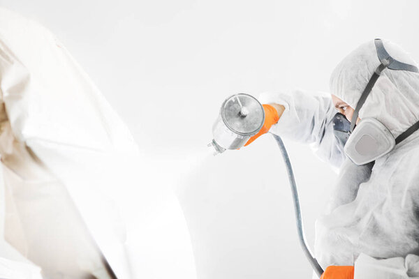 Body painter with airgun spraying white paint on a automobile in car maintenance service paint room. Worker sprays the vehicle with spray gun.