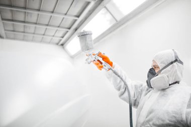 Odamda araba boyama elementlerini boyayan erkek bir tamirci. İşçi aracı sprey silahla beyaza boyuyor..