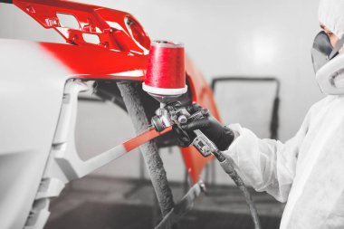 İşçi araba tamponunu sprey silahla boyuyor. Tamir sırasında boya odasındaki kırmızı araba elementleri.