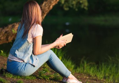 Gün batımında nehir kenarında kitap okuyan güzel bir kadın. Doğada sessizce bir kitap oku.