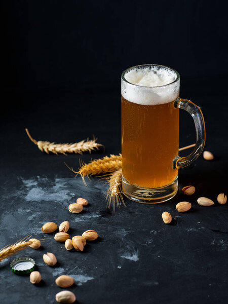 Стакан пшеничного пива на тёмном столе с пшеничными ушами и фисташками на закуску