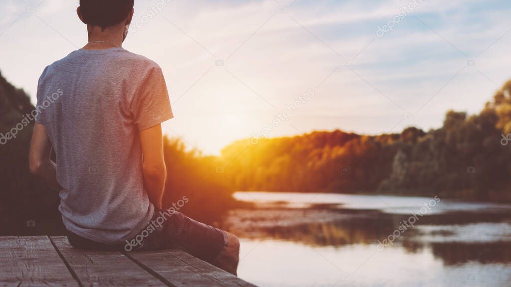 One guy sits on a wooden bridge by the river and admires the sunset and nature in the summer