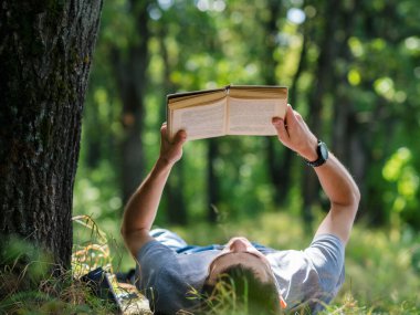 Genç adam parkta çimlerin üzerinde kitap okuyor.