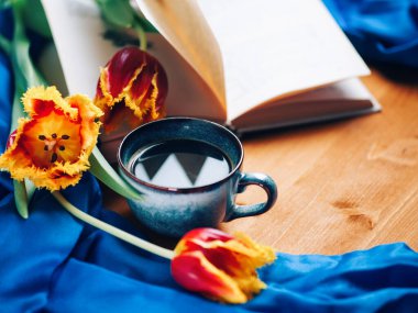 Bir fincan kahveyle bahar kompozisyonu, açık bir kitap ve tahta bir masada laleler.