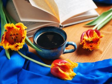 Bir fincan kahveyle bahar kompozisyonu, açık bir kitap ve tahta bir masada laleler.