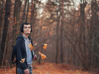 Genç yakışıklı bir adam sonbahar parkında yürüyor ve kulaklıkla müzik dinliyor ve sonbahar yapraklarını havaya fırlatıyor.