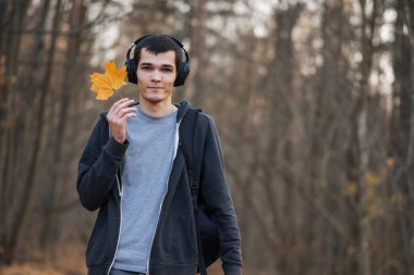 Genç yakışıklı bir adam sıcak bir akşamda sonbahar parkında yürüyor ve kulaklıkla müzik dinliyor.