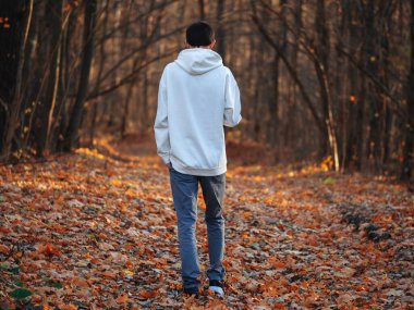 Beyaz kapüşonlu bir adam sonbahar ormanında yürüyor. Yolda bir sürü kuru sonbahar yaprağı var. Arka görünüm