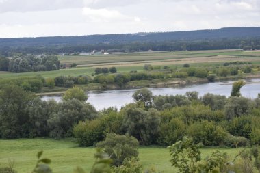 Tepenin tepesinden panoramik manzara yeşillik ve uzak ufuklarla çevrili bir nehri gösteriyor. Önplanda çalılıklar görünür, arkaplan biraz bulanık.