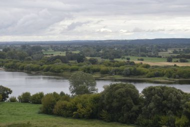 Tepenin tepesinden panoramik manzara yeşillik ve uzak ufuklarla çevrili bir nehri gösteriyor. Önplanda çalılıklar görünür, arkaplan biraz bulanık.