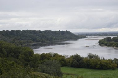 Yoğun ormanlar ve tepe arazili çalılarla çevrili bir nehrin panoramik manzarası ve arka planda parçalı bulutlu bir gökyüzü