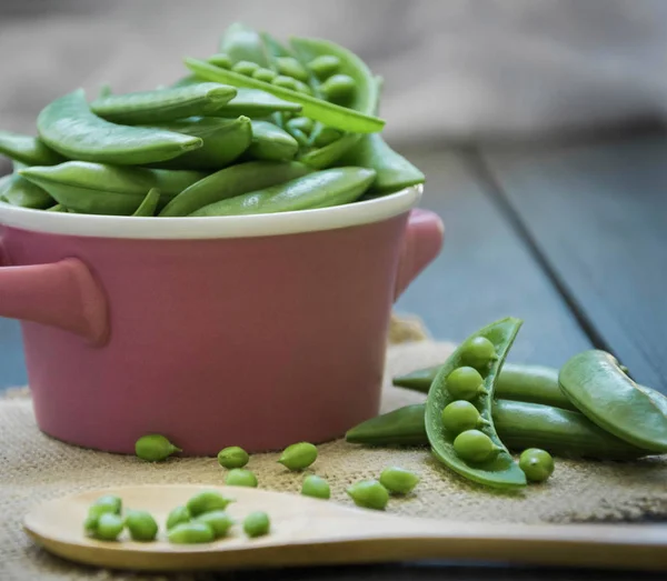 Pods of peas in a small pink saucepan, with a wooden spoon with peas, on a wooden board.In horizontal format.