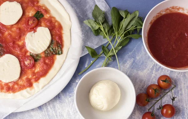 Üstten görünüm bir kase ezilmiş domates, yanındaki bir ham margarita pizza mozzarella peynir domates ve fesleğen birkaç yaprağı.