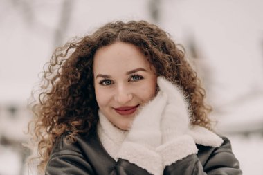 Kış sahnesinde kıvırcık saçlı bir kadın rüya gibi gülümsüyor. Deri ceket ve eldiven giyiyor, arka planda kar yağıyor. Görüntü kış modasını, güzelliği ve soğuk havanın neşesini ön plana çıkarıyor..